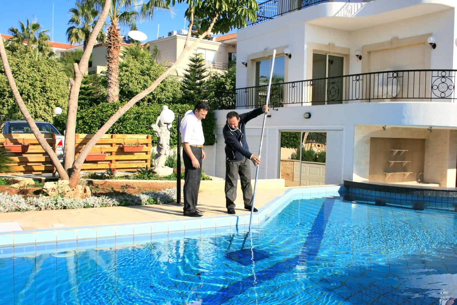 Dos hombres al lado de la piscina mirando el agua mientras uno de ellos la limpia con un limpiafondos.